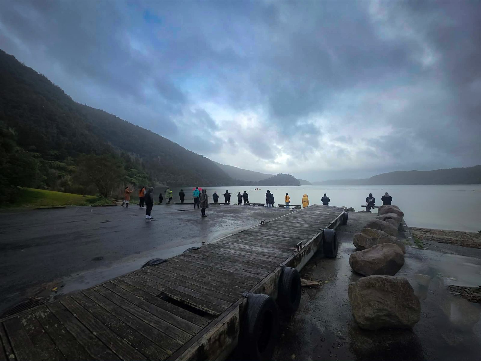 Whānau standing together along the lake edge at Okataina on an overcast morning, the maunga in mist across the water